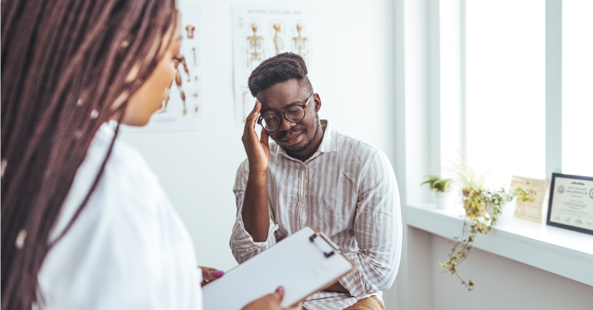 young man rubbing his forehead in agony while his doctor is writing out a prescription