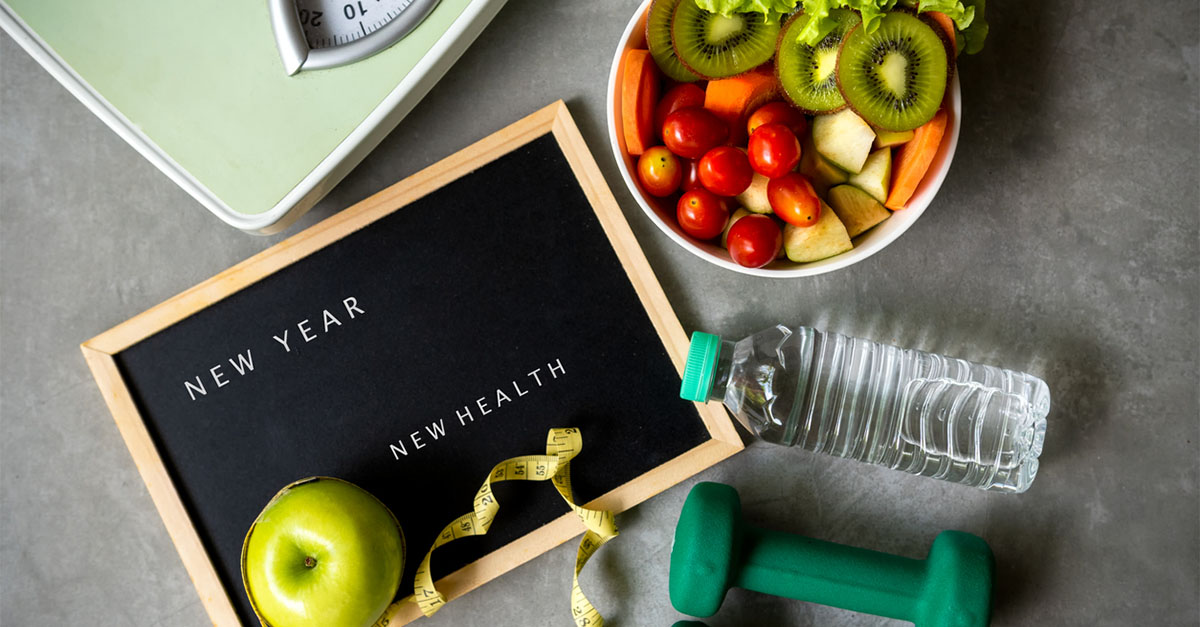 A sign that says “New Year, New Health,” a bowl a fruit, a water bottle, weights, measuring tape, and a scale.